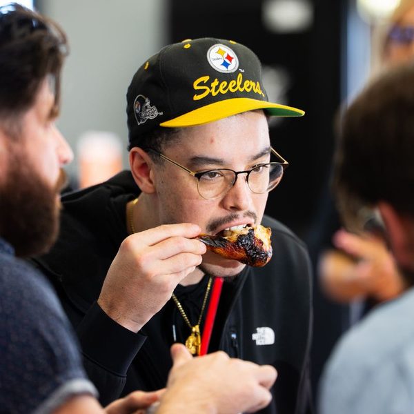 A customer in a Steelers cap and glasses biting into a charred chicken drumstick at a Sticky Chikky event, crowd visible behind him