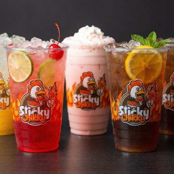 Five Sticky Chikky branded cups on a dark surface: cherry limeade with a lime wheel and cherry, a strawberry shortcake shake topped with whipped cream, iced tea with a lemon slice and fresh mint, and two more specialty drinks