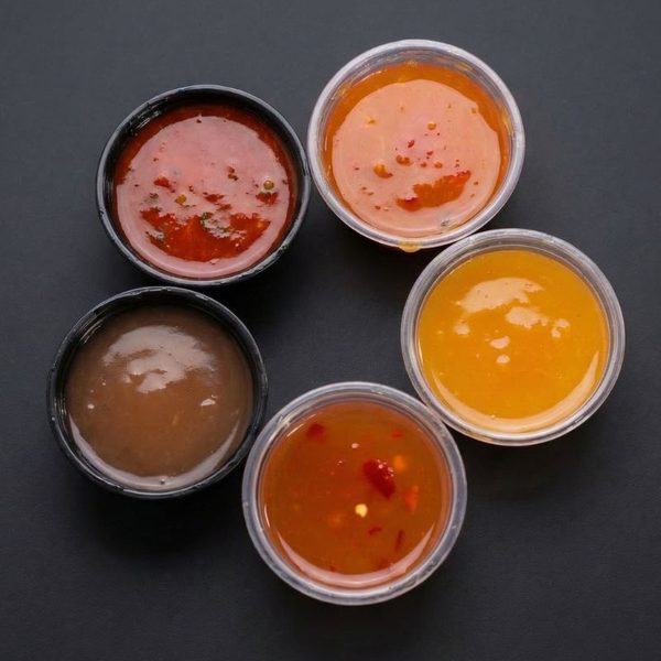 Five house-made sauce cups overhead on a dark grey surface: deep red, coral pink, golden yellow, brown, and translucent amber with chili flakes